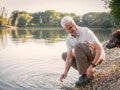 Homem de meia idade agachado à beira de um lago, sorrindo ao interagir com seu cachorro.