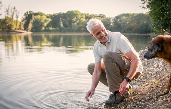 Homem de meia idade agachado à beira de um lago, sorrindo ao interagir com seu cachorro.